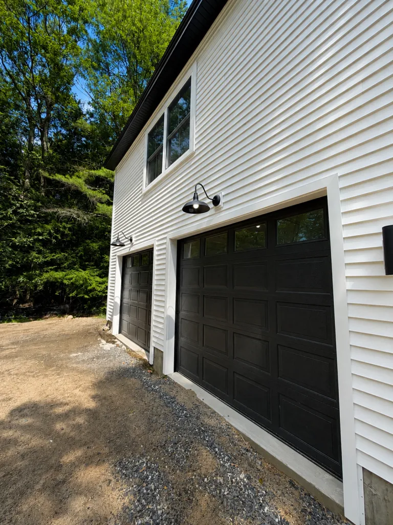 Detached garage with black overhead doors, barn lights, and white vinyl siding in Maine